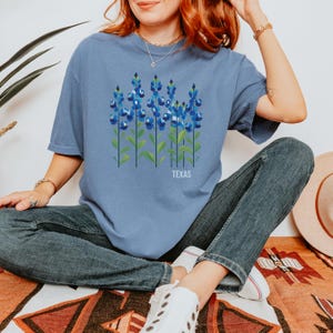 May include: A blue t-shirt with a graphic of bluebonnets, the Texas state flower, and the word "TEXAS" printed below the flowers.