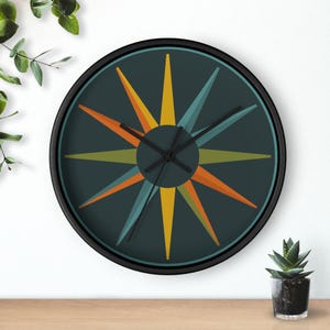 May include: Round wall clock with a black frame and a dark teal face. The clock features a colorful starburst design with orange, yellow, and teal points. The clock hands are black. A small potted succulent is visible in the lower right corner.