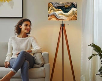 May include: A wooden tripod floor lamp with a rectangular lampshade. The lampshade features an abstract design in shades of brown, beige, gray, and teal. The lamp is positioned next to a woman sitting on a gray sofa.