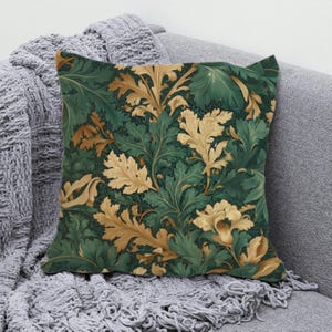 May include: Square throw pillow with a botanical pattern. The pillow features a repeating design of green and gold leaves on a dark green background. The pillow is resting on a gray sofa with a gray knitted throw blanket.