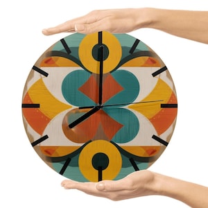 May include: A round wall clock with a colorful geometric design in orange, yellow, teal, and brown. The clock has black hands and black markers. The clock is held by two hands against a white background.