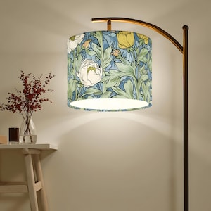 May include: A floor lamp with a floral patterned lampshade. The lampshade features a design of white and yellow flowers with green leaves on a blue background. The lamp has a curved, bronze-colored metal frame.