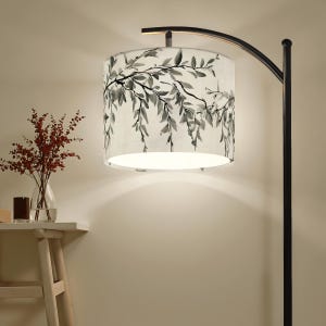 May include: A black arc floor lamp with a white lampshade featuring a black and gray botanical design. The lamp is positioned in a room with a neutral-colored wall. A small table with a vase of red flowers is in the background.