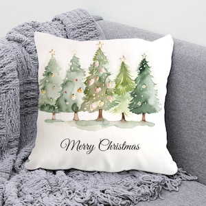 May include: White square pillow with a watercolor design of five Christmas trees in varying shades of green. Each tree is topped with a gold star and decorated with ornaments. The words "Merry Christmas" are written in black script.