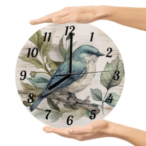 May include: Round wall clock featuring a blue bird perched on a branch, surrounded by green leaves. The clock face has black numerals and hands. The background has a wood grain pattern.