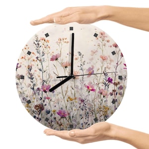 May include: A round wall clock featuring a floral design with pink, purple, and yellow flowers. The clock has black hands and square markers. The clock is held by two hands against a white background.