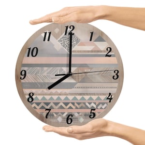 May include: A round wall clock with a light wood grain background and a geometric pattern in shades of gray, pink, and teal. The clock hands are black, and the numbers are black. The clock is held by two hands.
