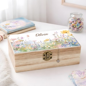 May include: A wooden keepsake box with a floral design on the lid, featuring the name "Olivia" in script. The box has a metal clasp and a heart-shaped pendant hanging from it. A jar of colorful items and a book are in the background.