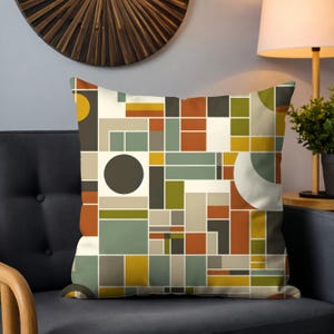 May include: Square throw pillow with a geometric pattern in shades of brown, green, yellow, and gray. The design features rectangles, squares, and circles. The pillow is on a dark gray couch.