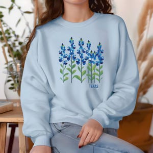 May include: Light blue sweatshirt with a graphic of bluebonnets, a Texas state flower, and the word "TEXAS" printed below the flowers.