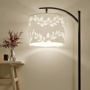 May include: A floor lamp with a black metal frame and a curved arm supporting a cylindrical lampshade. The lampshade is beige with a white leaf pattern. A small table with a vase of red flowers and a candle sits nearby.
