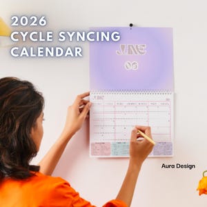May include: A 2026 Cycle Syncing Calendar is displayed on a white wall. The calendar is open to June, with a person writing on it with a pen. The calendar has a purple gradient background and the text "Aura Design".