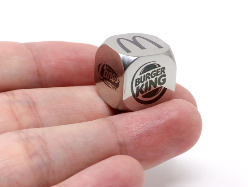 Pode incluir: Um dado de metal prateado com os logotipos do Burger King, McDonald's e Cane's em lados diferentes.