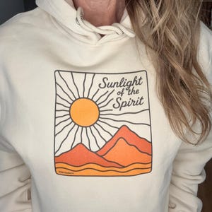 Peut inclure: Un sweat à capuche crème avec un motif graphique représentant un soleil, des montagnes et le texte "Sunlight of the Spirit". Le design incorpore des couleurs orange, jaune et noir. Le sweat à capuche a une capuche à cordon.