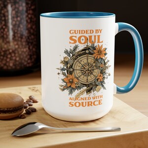 Pode incluir: Caneca de cerâmica branca com borda e alça turquesa. A caneca apresenta uma bússola com detalhes florais e o texto "Guided by Soul Aligned with Source".