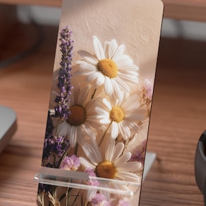 May include: A floral phone stand featuring a bouquet of white daisies with yellow centers, lavender, and pink flowers. The stand is propped up on a wooden desk, with a keyboard and headphones in the background. The design is printed on a rectangular stand.