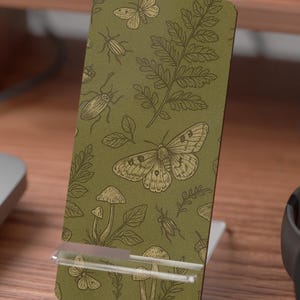 May include: A green phone stand with a nature-themed pattern featuring butterflies, beetles, mushrooms, and ferns. The stand is made of a sturdy material and is designed to hold a phone or tablet. The stand is on a wooden desk.