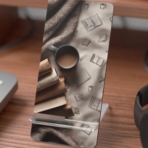 May include: A rectangular phone stand featuring a printed design of a stack of books, a dark mug filled with coffee, and a textured brown fabric. The background pattern consists of small line-drawn illustrations of open books and coffee cups.