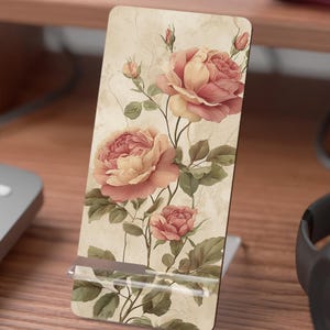 May include: A rectangular phone stand featuring a vintage floral design with pink and peach roses and green leaves. The stand has a neutral beige background and a white base. The stand is made of a durable material.