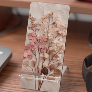 May include: A rectangular phone stand with a floral design. The stand features a beige background with pink and white flowers and brown leaves. The stand is made of a clear plastic material and is on a wooden desk.
