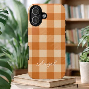 May include: A rectangular mobile phone case featuring an orange and cream gingham check pattern. The name Angel is printed in white cursive script on the lower portion of the case. The case has a black camera cutout with two circular lenses.