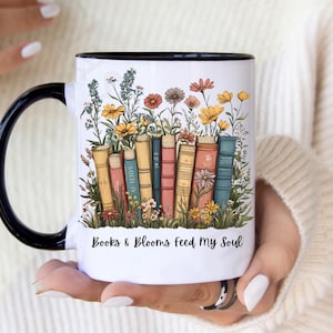 Ceramic mug with black handle featuring a colorful stack of books surrounded by blooming wildflowers and the phrase “Rooted in Stories, Blooming in Wisdom.”