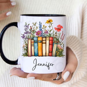 Personalized ceramic mug with black handle featuring a row of colorful books with blooming wildflowers and a custom name “Jennifer” in script – spiritual gift for readers and teachers.