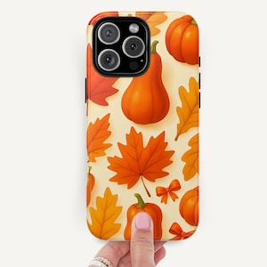 Könnte beinhalten: Eine Handyhülle mit einem herbstlichen Muster. Das Design zeigt orangefarbene Kürbisse, Ahornblätter und Schleifen auf cremefarbenem Hintergrund. Die Hülle wird von einer Hand mit Ringen gehalten.