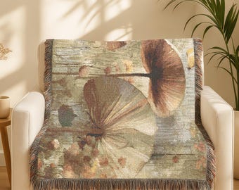 May include: A decorative throw blanket featuring a mushroom design in shades of brown, beige, and green. The blanket is draped over a white armchair, with a fringed edge. The design has a rustic, natural aesthetic.
