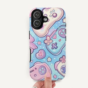 May include: A phone case with a pastel blue background featuring a pattern of video game controllers, hearts, stars, and flowers in shades of pink, purple, and white. The case is held by a hand.