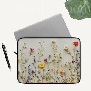 Puede incluir: Funda para portátil con un diseño floral de flores silvestres coloridas. La funda tiene una cremallera negra y un bolígrafo negro está al lado. El fondo es blanco con un portátil.