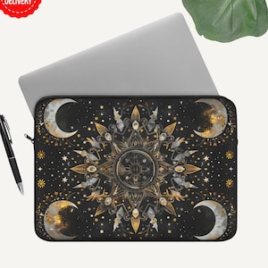 Puede incluir: Funda negra para portátil con un diseño celestial con un sol central, lunas y estrellas doradas y blancas. Un bolígrafo negro descansa junto a la funda. La funda está sobre un portátil gris claro.
