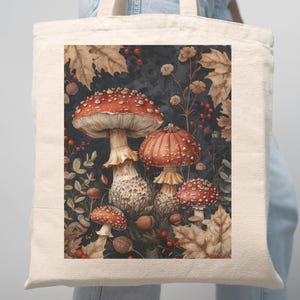 May include: A beige tote bag featuring a colorful illustration of red-capped mushrooms with white spots, surrounded by autumn leaves, berries, and nuts against a dark background. The bag has two beige handles.