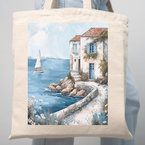 May include: A beige tote bag featuring a watercolor painting of a coastal scene. The artwork depicts a white building with blue shutters, a sailboat on the water, and a stone pathway. The scene is set against a blue sky and sea.