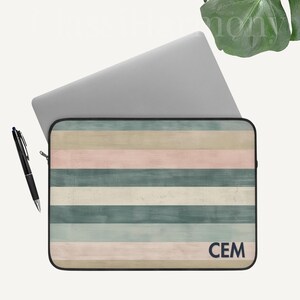 Puede incluir: Funda para portátil con un patrón de rayas pastel en tonos rosa, verde y beige. La funda tiene un borde negro y las iniciales "CEM" en la esquina inferior derecha. Un bolígrafo negro está al lado de la funda. El texto "Personalización Disponible" está en la esquina superior izquierda.