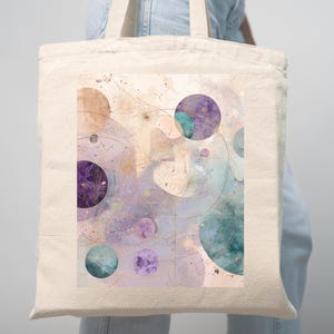 May include: Cream-colored tote bag featuring a watercolor design of overlapping circles in shades of purple, teal, and peach. The design includes gold lines and starburst details, creating an abstract celestial theme.