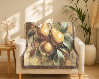 May include: A woven throw blanket featuring a detailed illustration of pears on a branch with green leaves. The color palette includes shades of yellow, green, and brown. The blanket is draped over a white armchair.