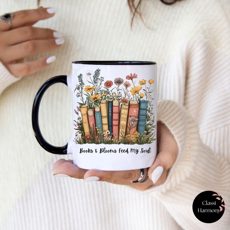 Ceramic mug with black handle featuring a colorful stack of books surrounded by blooming wildflowers and the phrase “Rooted in Stories, Blooming in Wisdom.”