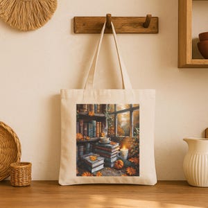 Cotton canvas tote bag featuring a cozy autumn library scene with stacked vintage books, a steaming coffee cup, glowing candle, pumpkins, and fall leaves by the window. Perfect gift for book lovers and cozy fall style