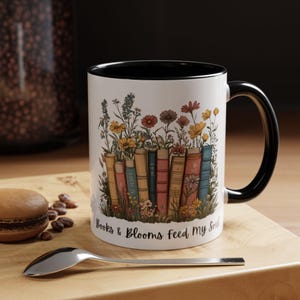 Ceramic mug with black handle featuring a colorful stack of books surrounded by blooming wildflowers and the phrase “Rooted in Stories, Blooming in Wisdom.”