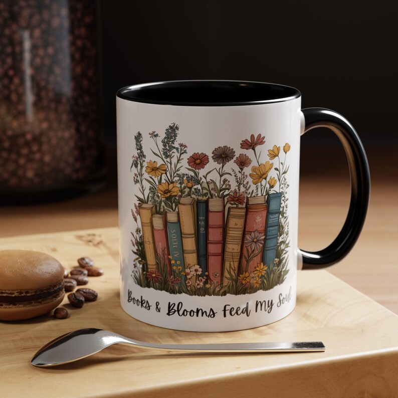 Ceramic mug with black handle featuring a colorful stack of books surrounded by blooming wildflowers and the phrase “Rooted in Stories, Blooming in Wisdom.”