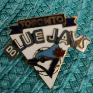 Può includere: Una spilla dei Toronto Blue Jays con un design bianco, blu e oro. La spilla presenta il nome e il logo della squadra, tra cui una palla da baseball e una foglia d'acero. La spilla ha la forma di un triangolo con uno stendardo in alto.