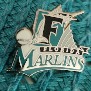 May include: A silver and teal Florida Marlins baseball pin featuring a marlin fish and a baseball. The pin reads "Florida Marlins" with a black "F" and a white baseball.