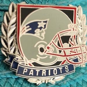 Könnte beinhalten: Eine silberne und rote Emaille-Anstecknadel mit einem Footballhelm mit einem blau-roten Patriots-Logo und dem Wort "PATRIOTS" auf einem blauen Banner. Der Helm ist von silbernen Lorbeerkränzen umgeben.