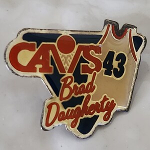 May include: A gold-colored pin with a blue and red Cleveland Cavaliers logo, a basketball, and a jersey with the number 43 and the name Brad Dougherty.