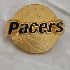 May include: A gold-colored pin featuring the word "Pacers" in black lettering. The pin has a basketball design in the background. The pin is round and has a slightly textured surface.