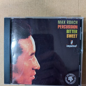 May include: A CD case with a black and white photo of Max Roach, a jazz musician, on the front. The title of the album is "Percussion Bitter Sweet" and the label is Impulse! Records. The back of the case features the GRP Presents Legendary Masters of Jazz logo.