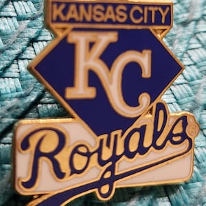 以下が含まれることがあります： カンザスシティ・ロイヤルズのロゴが入ったピンバッジ。ピンバッジは、白抜きの「KC」が入った青と白のひし形です。ひし形の上には、金色の長方形に「Kansas City」と金色の文字が入っています。ひし形の下には「Royals」の文字が金色で書かれています。