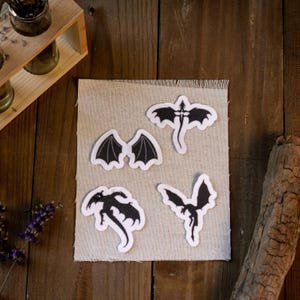 May include: A collection of five black dragon patches with white borders, displayed on a beige fabric square. The patches feature various dragon designs, including wings and full dragon silhouettes. The background is a wooden surface.
