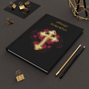 Pode incluir: Diário preto com uma cruz dourada e rosas vermelhas. O diário tem as palavras "always remember" impressas na capa.